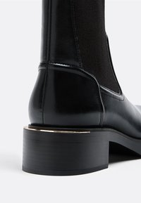 Czarny skórzany botek za kostkę z elastycznymi panelami bocznymi, okrągłym noskiem i grubym czarnym obcasem z złotym akcentem blisko podstawy.