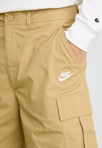 Mão no bolso de calças cargo bege da Nike com fecho de botão e logótipo branco bordado na coxa.