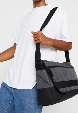Sac messager gris avec une base noire, texture en tissu et poche zippée à l'avant. Équipé d'une bandoulière noire réglable.