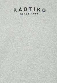 Tessuto grigio con testo ricamato nero "KAOTIKO SINCE 1999" centrato vicino alla parte superiore.