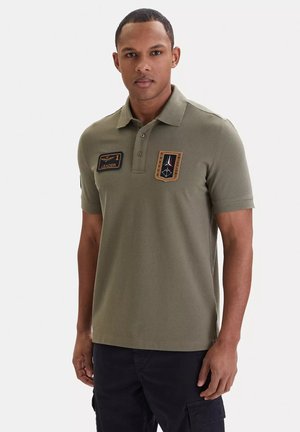 Uomo in piedi che indossa una polo verde oliva con toppe in stile militare e pantaloni cargo neri su sfondo neutro.