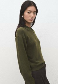 Suéter de mezcla de lana de color verde oscuro con cuello con solapa, tres botones y mangas largas. Textura suave, ajuste ligeramente holgado, usado con pantalones de cuadros.