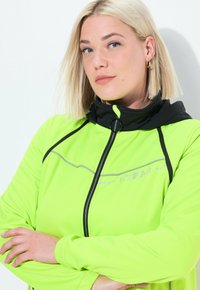 Neon vihreä takki mustalla hupulla, jossa on vetoketjusuljenta ja heijastavat yksityiskohdat. Sileä pinta, istuva muotoilu pitkillä hihoilla.