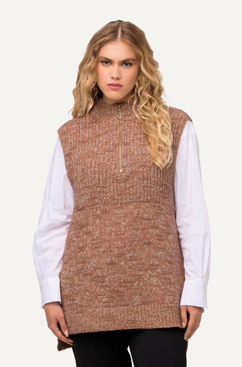 Gilet senza maniche in un filato marrone lavorato a maglia, con colletto alto e chiusura con mezza zip, indossato sopra una camicia bianca a maniche lunghe.