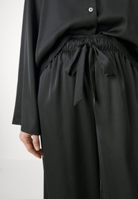 Svart satinblus med långa ärmar och knäppning framtill, med elastisk midja och snörning vid midjan för justerbar passform.