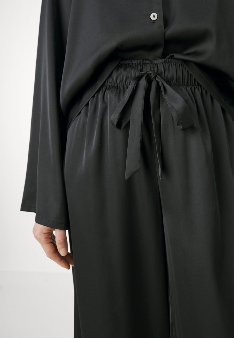 Blusa de satén negro con mangas largas y cierre de botones, que presenta una cinturilla elástica y un lazo en la cintura para un ajuste ajustable.