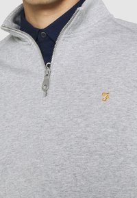 Sudadera gris con cremallera y cuello alto, que presenta un interior de cuello en color navy y un pequeño logotipo bordado en amarillo en el pecho. Textura suave.