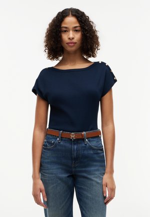 Žena s kučeravými vlasmi v tmavomodrom topu s gombíkmi na ramenách, hnedým opaskom a modrými džínsami, stojaca na bielom pozadí.