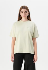 Young woman with long brown hair wears a light green oversized t-shirt and black pants, standing against a white background.