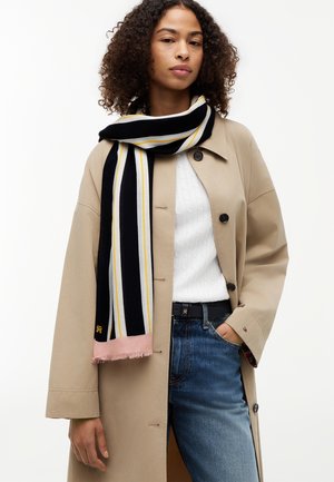Woman wearing beige trench coat, white textured sweater, blue jeans, and black scarf with white, yellow stripes, and pink fringe.
