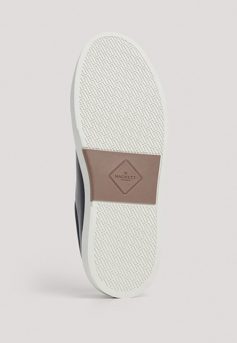 Semelle de chaussure blanche avec un motif texturé et une étiquette rectangulaire marron au centre affichant le logo "Hackett London".