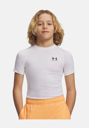 Blond kind draagt een aansluitend wit Under Armour T-shirt met korte mouwen en losse oranje sportshorts, staand met handen in de zakken.