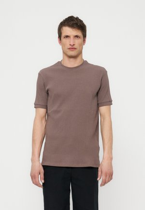 Mężczyzna z krótkimi brązowymi włosami, ubrany w brązową prążkowaną koszulkę z krótkim rękawem i czarne spodnie, stojący na tle jednolitego białego tła.