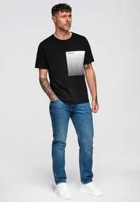 Černé bavlněné tričko s bílým pruhovaným panelem, na kterém je slovo "ombre". Nosené s modrými džíny a bílými teniskami. Jednoduché pozadí.