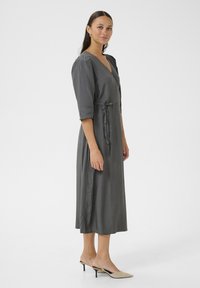 Gray wrap dress with a V-neck, short sleeves, and a tie waist. Made of smooth fabric, it falls to mid-calf, paired with light-colored heels.