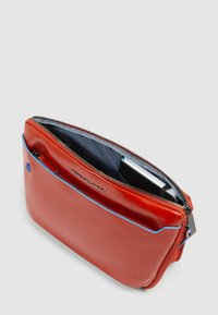 Borsa in pelle rossa con una texture liscia, chiusura con zip, dettagli blu e un scomparto interno visibile, progettata per contenere un quaderno e piccoli oggetti.