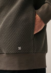 Felpa grigio scuro con texture, caratterizzata da un motivo trapuntato, polsini a costine e un piccolo logo argentato vicino all'orlo.