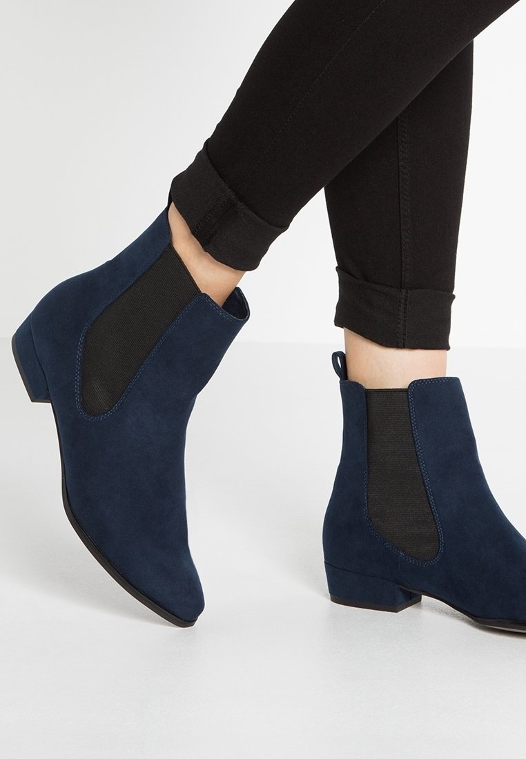 Bottines en daim bleu marine avec panneaux latéraux élastiques noirs portées avec un pantalon skinny noir retroussé sur fond clair.