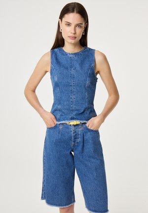 Vrouw met een mouwloos blauw denim topje en bijpassende knielange denim shorts met een gele riem, staand met haar handen in de zakken.