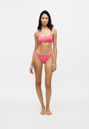 Femme debout pieds nus portant un soutien-gorge de sport rose Nike et un bas de bikini assorti sur un fond blanc uni.