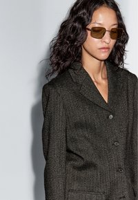 Kobieta z kręconymi włosami do ramion, nosząca prostokątne brązowe okulary przeciwsłoneczne i ciemną teksturowaną marynarkę, spojrzana nieco w dół na jasnym tle.