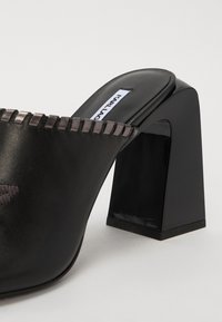 Crne klompe s potpeticama koje imaju glatki kožni gornji dio, blok petu i ušivenu detalj uz rub otvora.
