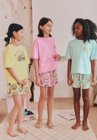 Kolm tüdrukut kannavad värvilisi puuvillaseid T-särke ja sobivaid lühikesi pükse, millel on multikatoidumustrid. T-särkide värvid on kollane, roosa ja mündiroheline.