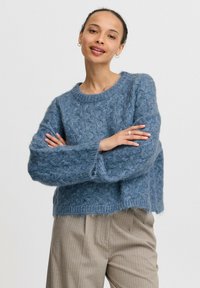 Sininen neulepaita, jossa on tekstuuriompelemallia, pyöreä pääntie ja leveät hihat. Yhdistetty beigen raitahousujen kanssa.