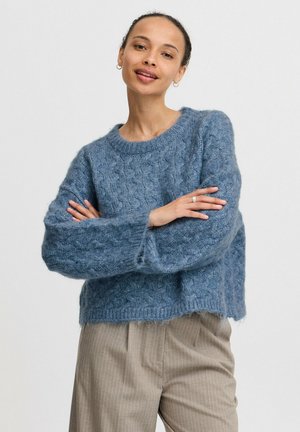 Pull en tricot bleu avec un motif de câble texturé, col rond et manches larges. Associé à un pantalon beiges à rayures.