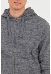 Sudadera gris con cremallera, hecha de tela texturizada, que cuenta con capucha, cordones y bolsillos frontales. Cierre frontal con cremallera.