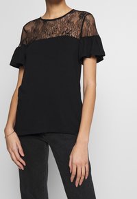 Kvinna i svart topp med genomskinlig blommig spets, puffärmar och mörka jeans, samt ett tunt armband på vänster handled.