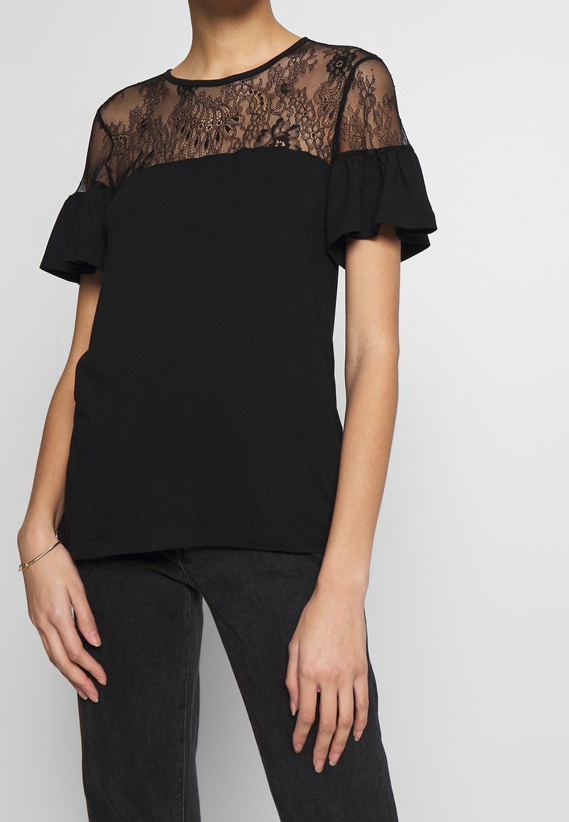 Kvinna i svart topp med genomskinlig blommig spets, puffärmar och mörka jeans, samt ett tunt armband på vänster handled.