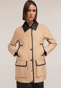 Beige nepromokavá bunda s černým límcem, zdobená zlatými knoflíky, dvěma předními kapsami a černým lemem podél okrajů.