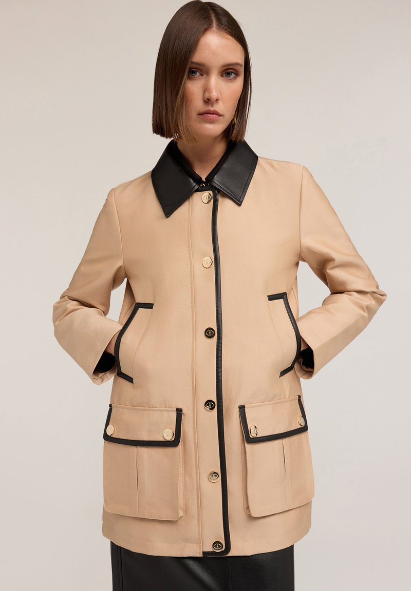 Beige nepromokavá bunda s černým límcem, zdobená zlatými knoflíky, dvěma předními kapsami a černým lemem podél okrajů.
