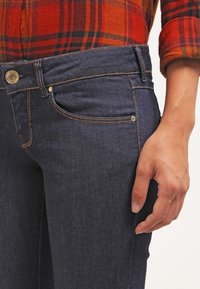 Dunkelblaue Jeans aus Denim mit mittelhohem Bund, kontrastierenden Nähten, Vordertaschen und einem metallischen Markenknopf. Hand ruht an der Seite.