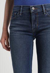 Dark blue denim jeans with a fitted design, featuring five pockets, visible stitching, and a metal button closure at the waist.
