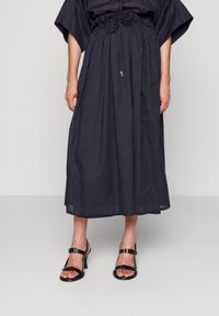 Nainen, joka pukeutuu pitkään, tummansiniseen hameeseen, jossa on joustava, sidottu vyötärö ja mustat, remmillä varustetut korkokengät, seisoo yksinkertaista taustaa vasten.