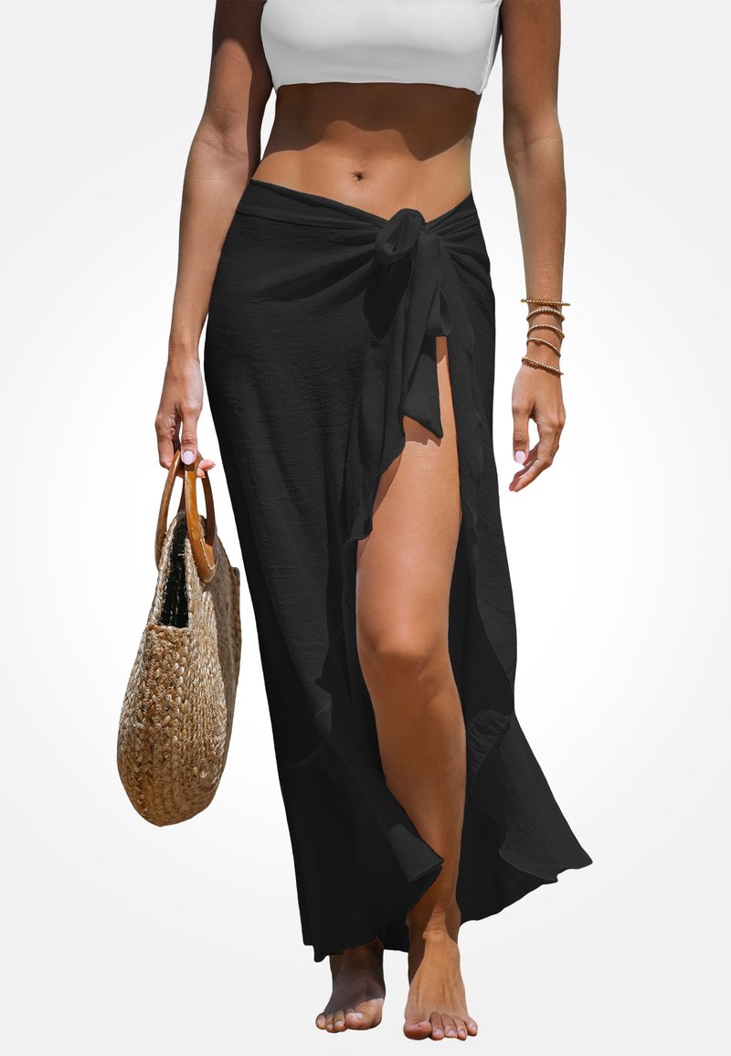 Falda negra envolvente hecha de un tejido ligero, con un lazo en la cintura y una alta abertura, complementada con un bolso de mimbre y pies descalzos.