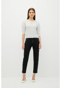Hellgrauer gestrickter Pullover mit Puffärmeln und einem strukturierten Muster, kombiniert mit hoch geschnittenen schwarzen Jeans. Weiße spitz zulaufende Absätze runden das Outfit ab.