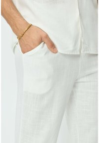 Pantalons en lin blanc avec un motif texturé, dotés d'une poche et d'une main reposant sur le côté, complétés par un bracelet en chaîne doré.
