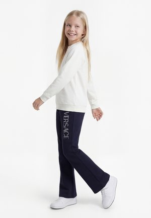 Usměvavá dívka s dlouhými blond vlasy, která kráčí, má na sobě bílou mikinu, tmavě modré kalhoty s nápisem "VERSACE" a bílé tenisky na bílém pozadí.