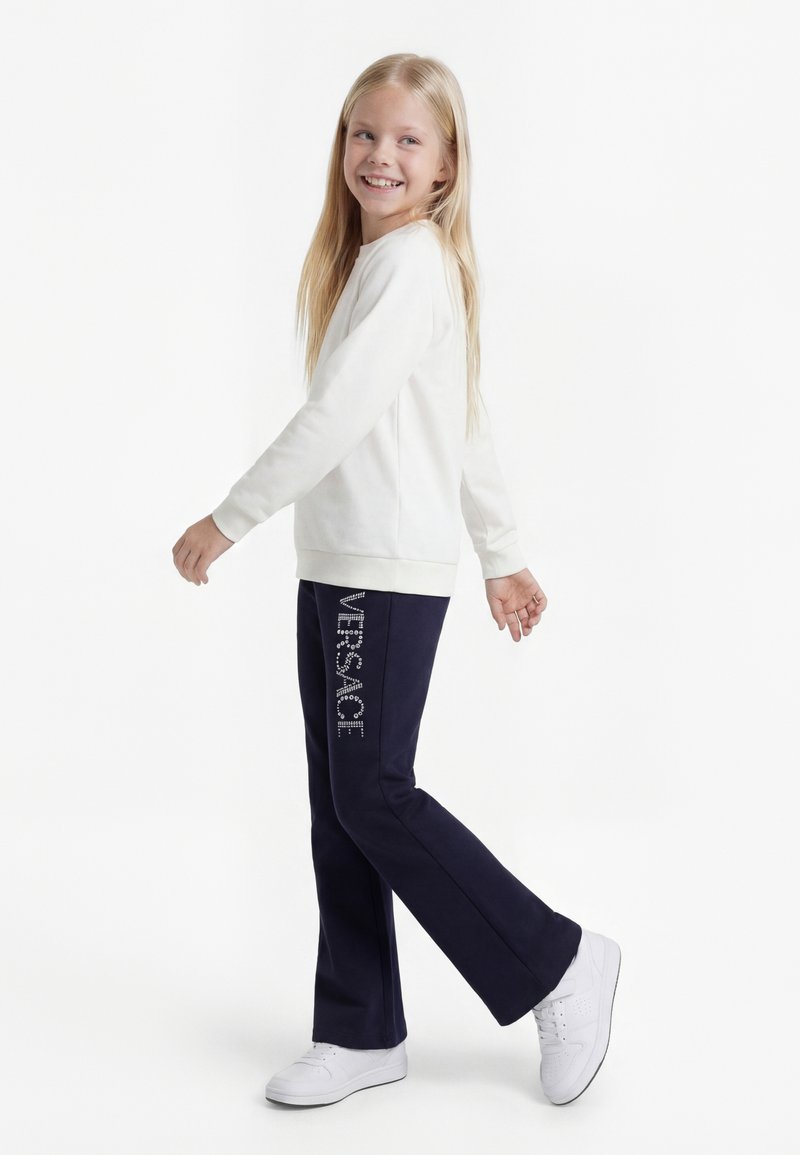 Niña sonriente con cabello largo y rubio caminando, lleva sudadera blanca, pantalones azul marino con el texto "VERSACE" y zapatillas blancas sobre fondo blanco.