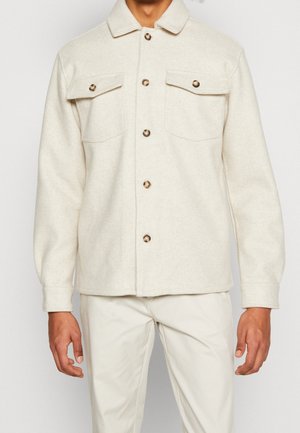 Veste beige en laine boutonnée avec deux poches à rabat sur la poitrine et pantalon beige assorti porté par une personne sur fond blanc.