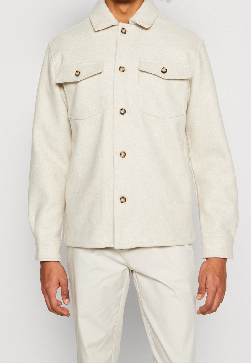Veste beige en laine boutonnée avec deux poches à rabat sur la poitrine et pantalon beige assorti porté par une personne sur fond blanc.