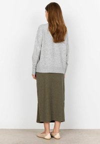 Šedý svetr přes midi sukni se zelenými pruhy, která má postranní rozparky. Modelka má na sobě béžové, vzorované boty na klínu a stojí před světlou zdí.