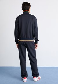 Pessoa em pé, de costas, vestindo um fato de treino preto com recortes vermelho, amarelo e verde, e ténis Adidas brancos com riscas vermelhas.