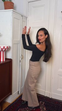 Femme aux longs cheveux foncés portant un top noir décolleté et un pantalon beige qui applaudit dans une pièce avec des portes blanches et des meubles en bois.