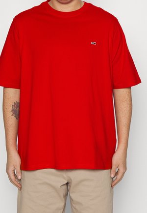 Osoba ubrana w prozną czerwoną koszulkę z krótkim rękawem z małym logo na piersi oraz beżowe spodnie, na lewym przedramieniu widoczny tatuaż.