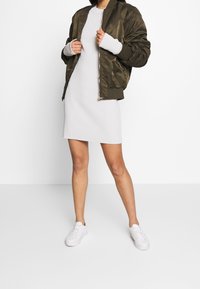 Olivgrüne Bomberjacke über einem figurbetonten, gerippten weißen Kleid; kombiniert mit weißen Sneakers. Glatte Textur mit Reißverschluss und gerippten Ärmeln.