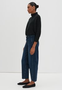 Maglione nero a trama con piccolo collo alto, abbinato a jeans in denim blu a vita alta. Scarpe nere basse completano l'outfit.